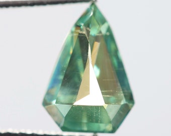 May include: A green pear-shaped gemstone with a faceted cut. The gemstone is held in tweezers and is illuminated by a light source.