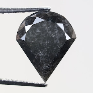 May include: A black diamond with a heart shape cut. The diamond is held by tweezers and has a rough, textured surface.