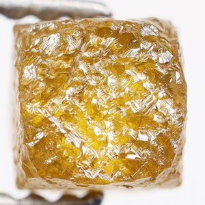 Puede incluir: Un diamante en bruto de color amarillo dorado con forma de cubo. La superficie de la gema es muy texturizada y refleja la luz, creando un efecto brillante. El diamante está sujeto por unas pinzas metálicas sobre un fondo blanco.
