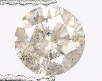 May include: A round, faceted diamond with a light gray hue. The diamond is held by a silver metal tool. The gem has a complex internal structure with visible inclusions, adding to its unique character. The facets reflect light, creating a sparkling effect.