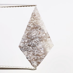 Puede incluir: Un diamante en bruto en forma de cometa, sujeto con unas pinzas. El diamante tiene una superficie texturizada y cristalina con tonos grises y marrones. La gema se encuentra sobre un fondo blanco.
