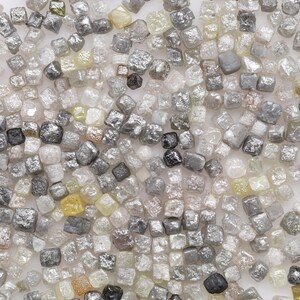 May include: A close-up view of a collection of small, rough, grey diamonds. The diamonds are various shades of grey and some have a slight yellow tint.