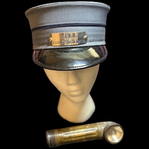 May include: A grey railroad cap with a black patent leather brim and a gold-toned metal badge reading L.I.R.R. ASSISTANT STATION MASTER, accompanied by a cylindrical, weathered brass-colored metal flashlight with a side-facing circular lens.