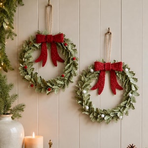 May include: Two circular wreaths made of green leaves hanging on a wall. Each wreath features a large, textured red bow at the top. One wreath is decorated with small red berries, while the other is adorned with small white pom-poms.