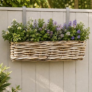May include: A rectangular woven wicker planter filled with green foliage and purple and white flowers, hanging from a grey fence. The planter is attached with grey metal hangers. The background shows a blurred view of green trees.
