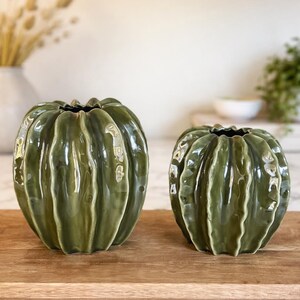 May include: Two green ceramic vases, pumpkin-shaped with a ribbed design, sit on a wooden tray. The vases have a glossy finish and dark openings. The background is blurred, with hints of a white wall and greenery.