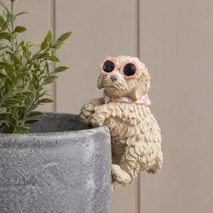 May include: A beige dog figurine wearing pink sunglasses and a pink and white scarf, perched on the edge of a gray plant pot. The dog is looking forward, with its paws resting on the pot's rim. Green plant leaves are visible.