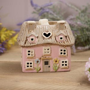 May include: A pink ceramic house-shaped candle holder with a thatched roof. The house features small windows, a heart-shaped opening, and floral accents. The candle holder is designed to hold a tea light.