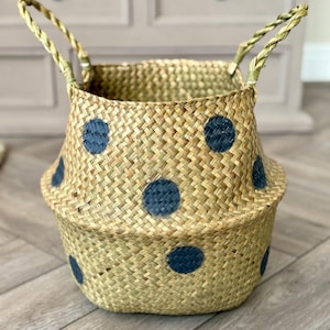 May include: Woven basket with a natural beige color and navy blue polka dots. The basket has two woven handles and a flared shape. This decorative basket is suitable for storage or as a decorative accent.