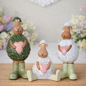 May include: Three ceramic sheep figurines with brown faces, holding pink hearts. One sheep is green with a textured pattern, another is white with a textured pattern, and the third is white. All wear green wellington boots and sit on a wooden surface.
