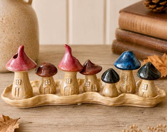 May include: A set of seven ceramic mushroom-shaped houses in various colors including red, brown, blue, and black, each featuring a small door and window, arranged on a long, wavy, light-colored ceramic tray with a textured, organic finish.