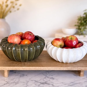 May include: Two decorative ceramic bowls filled with red apples. One bowl is dark green, the other is white. Both bowls have a ribbed design and sit on a wooden tray. The tray is on a marble surface.
