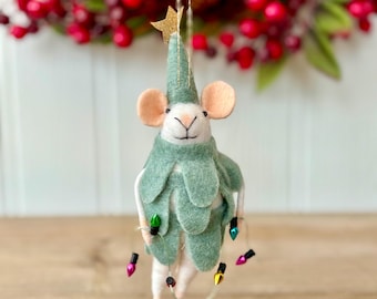 May include: A felt mouse ornament wearing a sage green pointed hat with a gold star and a matching leaf-shaped cape. The mouse has pink ears and holds a thin string of small, multi-colored decorative Christmas lights around its body.