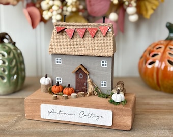 May include: A wooden autumn cottage decoration with a gray exterior, burlap roof, and red pennant banner. The base features miniature pumpkins, a broom, and the text "Autumn Cottage". Decorative pumpkins and foliage are in the background.