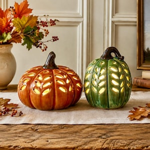 May include: Two ceramic pumpkin decorations, one orange and one green, featuring cut-out leaf patterns that allow internal light to shine through. Each pumpkin has a dark brown stem and a glossy finish, sitting on a light-coloured fabric runner.
