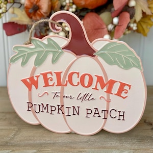May include: A pumpkin-shaped welcome sign with the words "WELCOME To our little PUMPKIN PATCH" in orange and brown. The sign has a light pink and white colour scheme with green leaves and a brown stem. A jute rope is attached for hanging.