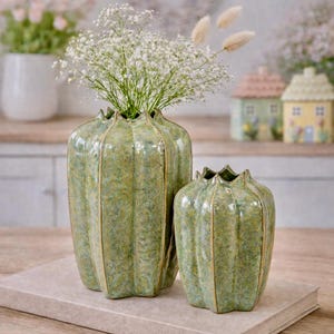 May include: Two green ceramic vases with a textured finish, one large and one small, sit on a neutral-toned book. The vases have a fluted design and hold white flowers. Decorative ceramic houses are in the background.