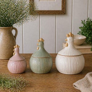 May include: Three ceramic hen-shaped jars in pink, sage green, and off-white. Each jar has a ribbed design and a decorative comb. The jars are displayed on a wooden surface. The jars are in different sizes.