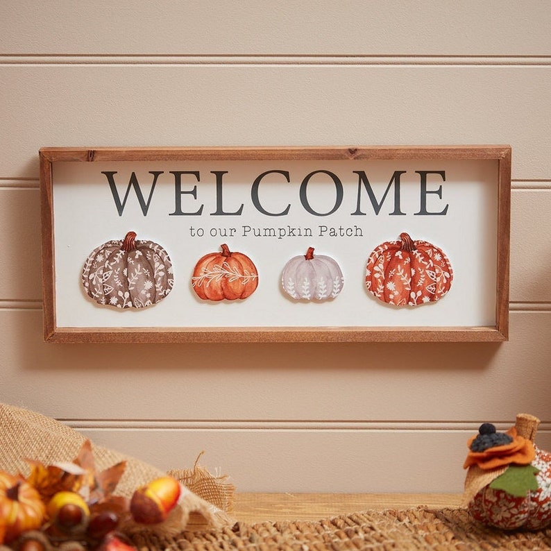 May include: A rectangular wooden framed sign with a white background featuring the black text WELCOME to our Pumpkin Patch. Below the text are four illustrated pumpkins in shades of brown, orange, and light grey, each with white leaf patterns.