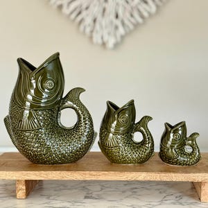 May include: Three green ceramic fish-shaped jugs of varying sizes are displayed on a wooden tray. The jugs have open mouths and detailed scale patterns. The largest jug is on the left, followed by a medium and a small one.