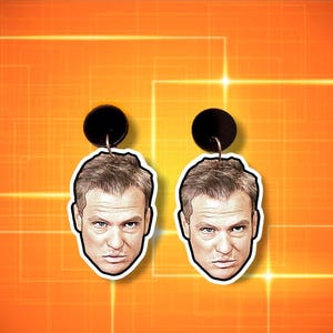 May include: A pair of novelty earrings with a man's face. The earrings have a black circular top and a white outline around the face. The background is orange with a grid pattern.