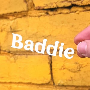 以下が含まれることがあります： 太字の白いサンセリフ体の大文字と小文字で「Baddie」という単語が印刷された、透明な長方形の粘着デカール。ステッカーは透明で、素材を通して背後の表面が見えるようになっています。