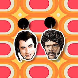 May include: A pair of novelty earrings featuring cartoon portraits of two men. One earring depicts a man with black hair and a cigarette, the other a man with an afro. The earrings have black circular tops and silver hooks.