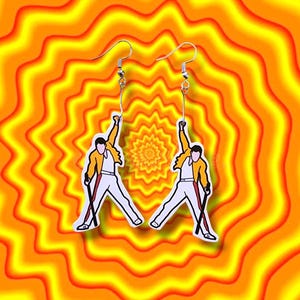 Puede incluir: Un par de pendientes de plata con una imagen de dibujos animados de una persona con una chaqueta amarilla y pantalones blancos sosteniendo un micrófono. Los pendientes están sobre un fondo de círculos concéntricos naranja y amarillo.