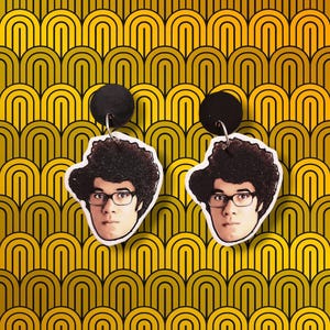 May include: A pair of earrings featuring a printed image of a man with curly brown hair and glasses. The earrings are made of black and white polymer clay and have a black metal hook.