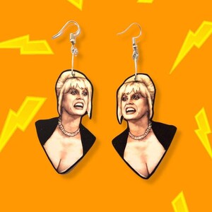 May include: A pair of dangle earrings featuring a woman's face with blonde hair and a black top. The earrings are silver and have a hook closure.