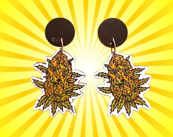 May include: A pair of earrings featuring a cannabis bud design. The earrings have black circular studs with gold-colored hooks. The cannabis bud is depicted in shades of green, yellow, and orange, with a white outline. The background is a bright yellow with radiating lines.