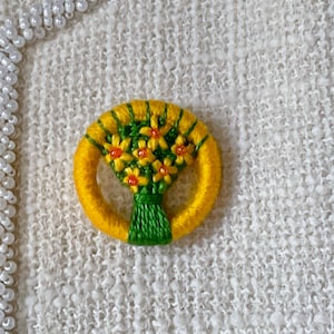 May include: A yellow button with a green embroidered flower design. The flowers are yellow with orange centres.