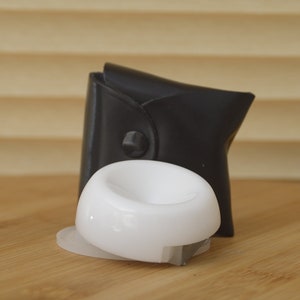 May include: A white plastic ring holder with a black leather case. The ring holder has a smooth, rounded surface and a small, flat base.