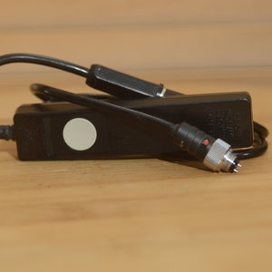 May include: A black remote control shutter release with a white button and a silver connector. The cable is black and coiled.