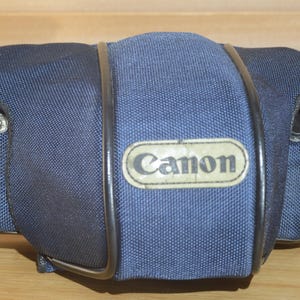 May include: A navy blue Canon camera case with a black trim and a metal frame. The case has a rectangular shape with rounded corners and a Canon logo on the front. The case is made of a textured fabric.