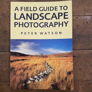 May include: A field guide to landscape photography book cover featuring a photo of a stone wall in a field with a building in the distance.