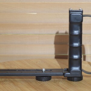May include: Black camera grip with a strap and a mounting plate. The grip has a textured surface and a screw-on base.