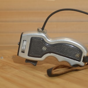 May include: A silver and black remote control shutter release with a black leather strap. The shutter release has a textured grip and a dial on the side.