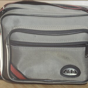 Puede incluir: Bolso de hombro de lona gris con ribetes negros y rojos. El bolso tiene un bolsillo con cremallera en la parte delantera y un logotipo plateado que dice "JRD".