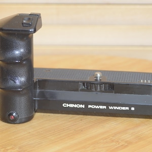 Puede incluir: Accesorio de cámara de película Chinon Power Winder S negro con un agarre texturizado y un botón plateado.