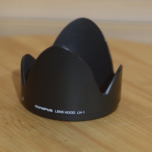Op de afbeelding: Zwarte Olympus lenskap LH-1. De lenskap is gemaakt van matzwart materiaal en heeft een tulpvormig ontwerp. De tekst "OLYMPUS LENS HOOD LH-1" staat in het wit op de zijkant. Ontworpen voor een cameralens.
