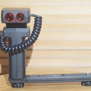 May include: A black camera flash with a coiled cord and two red lights. The flash has a mounting bracket on the bottom.