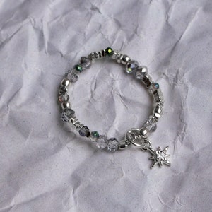 Puede incluir: Un brazalete plateado con un dije de estrella. El brazalete está hecho de pequeñas cuentas transparentes con acentos iridiscentes.