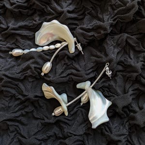 Puede incluir: Un par de pendientes únicos con elementos iridiscentes en forma de concha y perlas colgantes. Los pendientes tienen un diseño de metal plateado y están colocados sobre un fondo oscuro y texturizado.