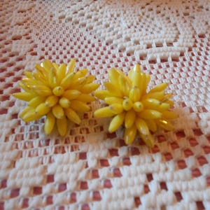 May include: Two yellow flower-shaped earrings with a cluster of small, oblong beads. The earrings are set against a white crocheted background with a floral pattern. The earrings are a bright, sunny yellow.