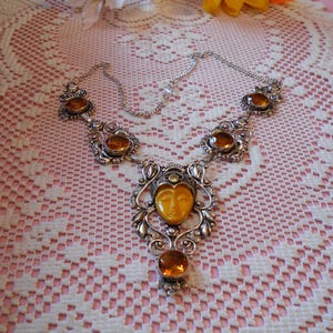 May include: A silver-toned necklace with amber-colored stones. The necklace features a central pendant with a carved face and a series of smaller stones. The necklace is displayed on a lace tablecloth.