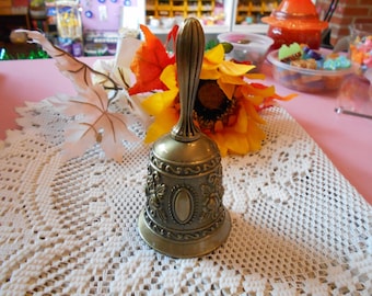May include: A vintage brass bell with an ornate floral design. The bell has a long handle and a detailed pattern around the base. It sits on a white lace tablecloth, with colorful flowers in the background. A decorative item.