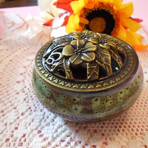 May include: A decorative ceramic jar with a bronze floral lid. The jar has a speckled green and brown glaze. The lid features a floral design. The jar is sitting on a white lace doily, with a sunflower in the background.