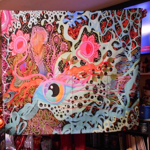 May include: A colorful tapestry with a psychedelic design. The artwork features an eye, organic shapes, and vibrant colors including pink, orange, blue, and black. The overall theme is abstract and artistic, suitable for wall decor.