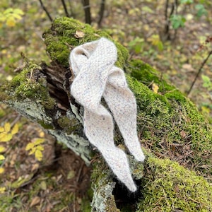 以下が含まれることがあります： 森の中に、苔むした木の切り株の上に、クリーム色の柔らかなニットのストールがかけられています。ストールには、かすかに斑点模様が入っています。